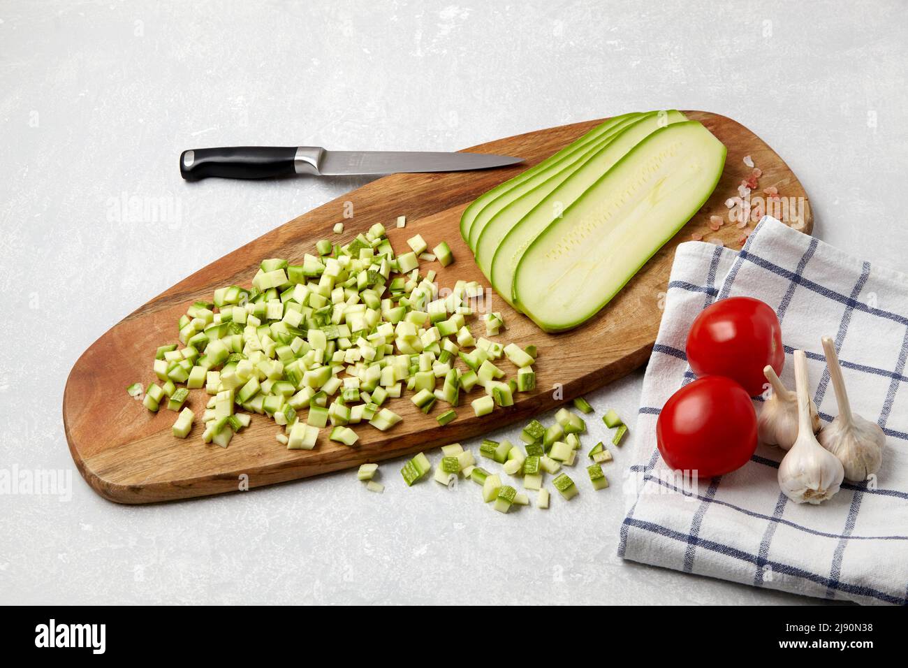 Affettare le zucchine di zucchine su un tagliere di legno, coltello, pomodori, aglio e tovagliolo su una tavola leggera in cemento Foto Stock