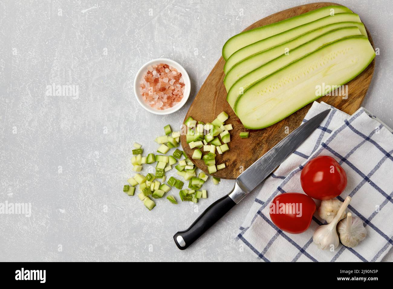 Affettare le zucchine di zucchine su un tagliere di legno, coltello, pomodori, aglio, sale rosa e tovagliolo su un tavolo in cemento leggero. Vista dall'alto piatta Foto Stock