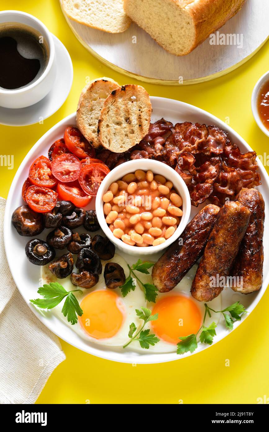Colazione all'inglese completa con uova fritte, pancetta, salsiccia, fagioli e funghi su sfondo giallo. Foto Stock