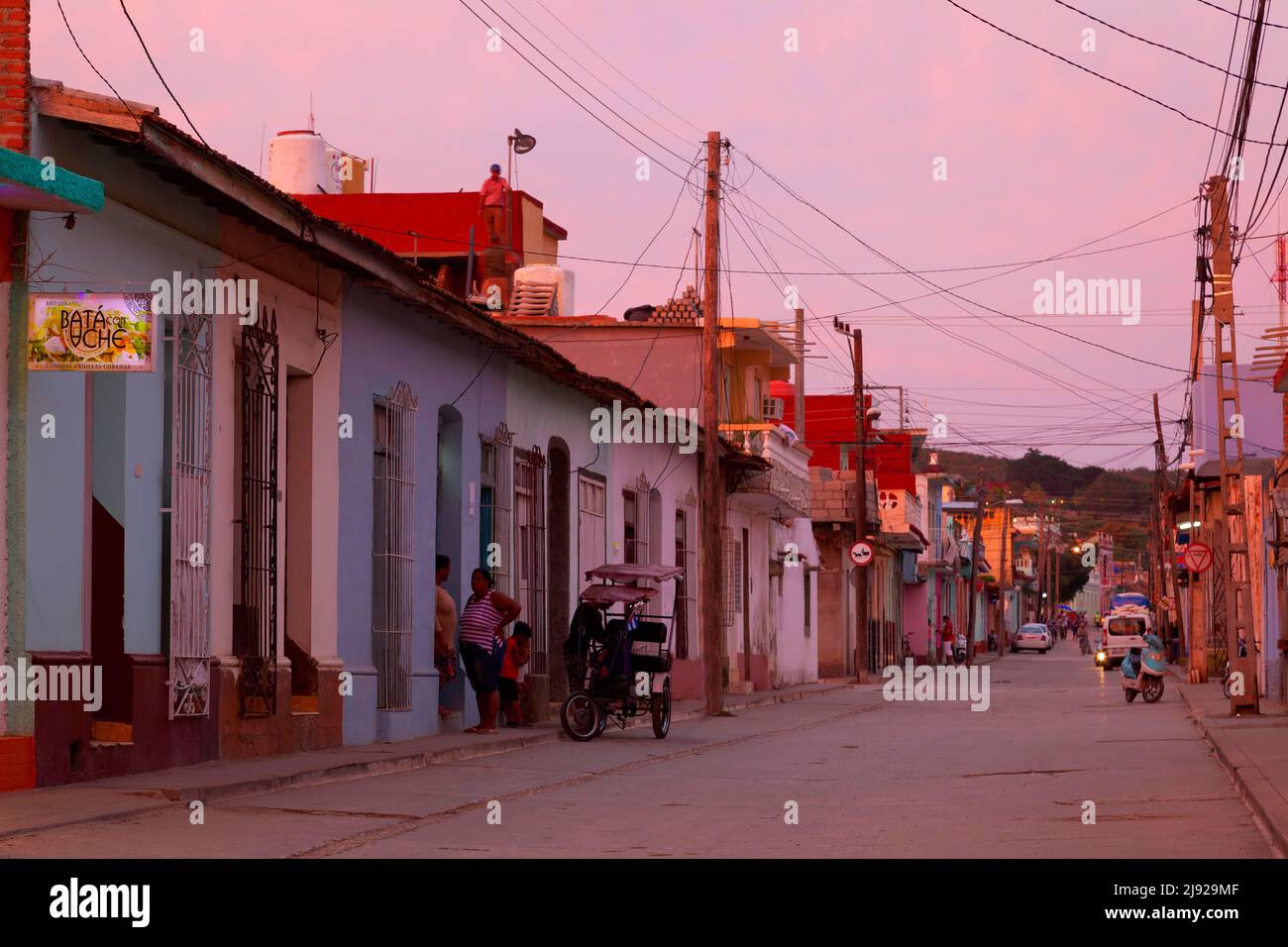Pittoresca scena di strada dopo il tramonto, ciottoli, telegrafo palo con linee elettriche, persone, cubani in piedi di fronte alla porta d'ingresso, auto, moto Foto Stock
