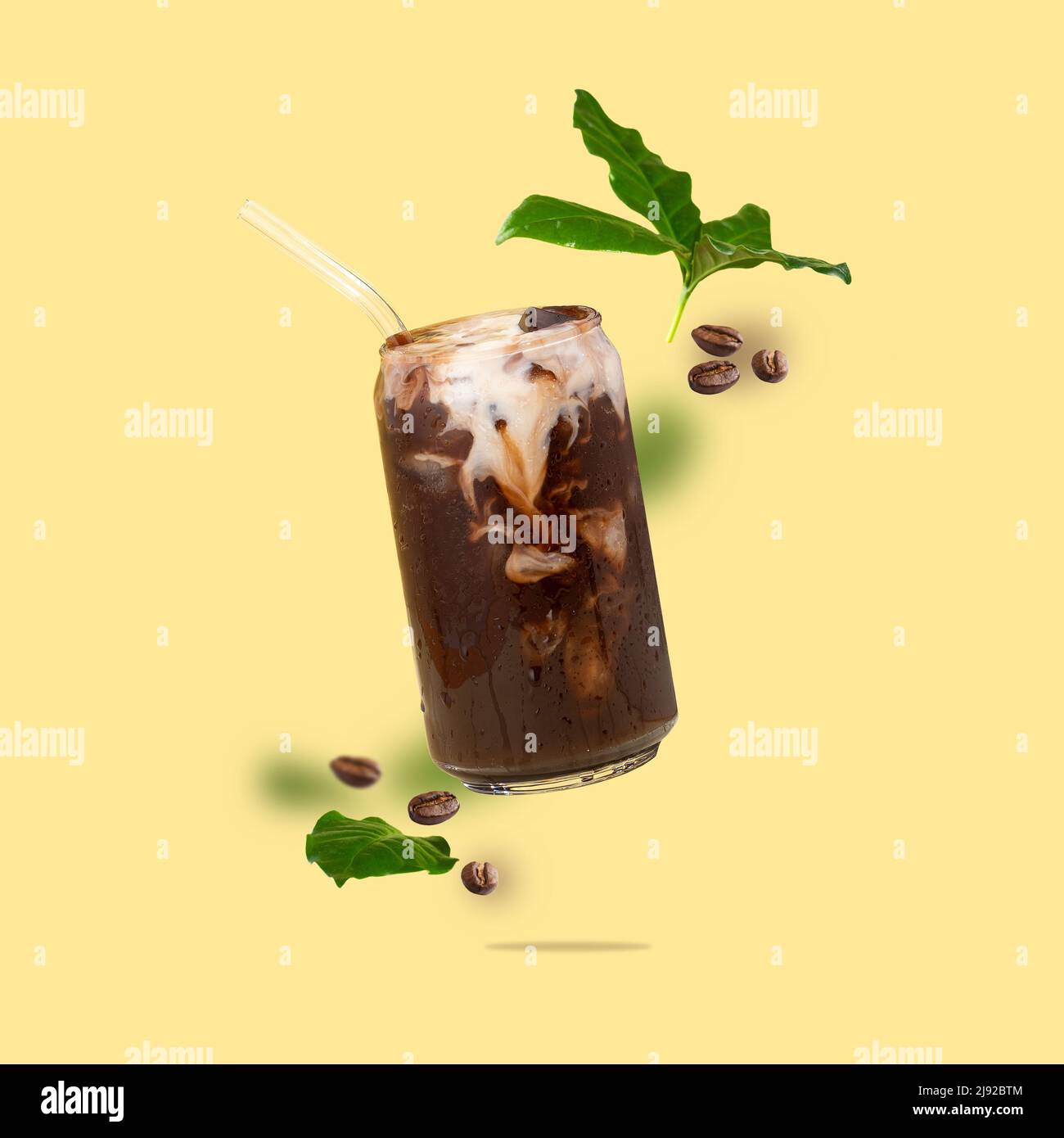 Tazza galleggiante in vetro a forma di lattina con caffè, chicchi di caffè, foglie di caffè che volano su sfondo giallo. Bevanda di caffè con latte vegetale. Foto Stock