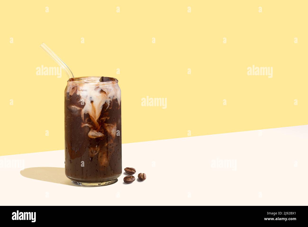 Caffè in chicchi e bevanda fredda con cannuccia di vetro su sfondo di proiezione diagonale isometrica giallo. Bicchiere di caffè a forma di lattiera a base di piante Foto Stock