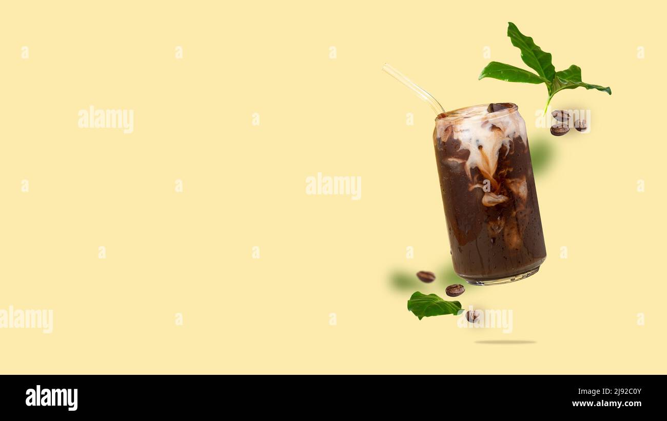 Tazza galleggiante in vetro a forma di lattina con caffè, chicchi di caffè, foglie di caffè che volano su sfondo giallo bevanda a base di caffè con latte vegetale. Foto Stock