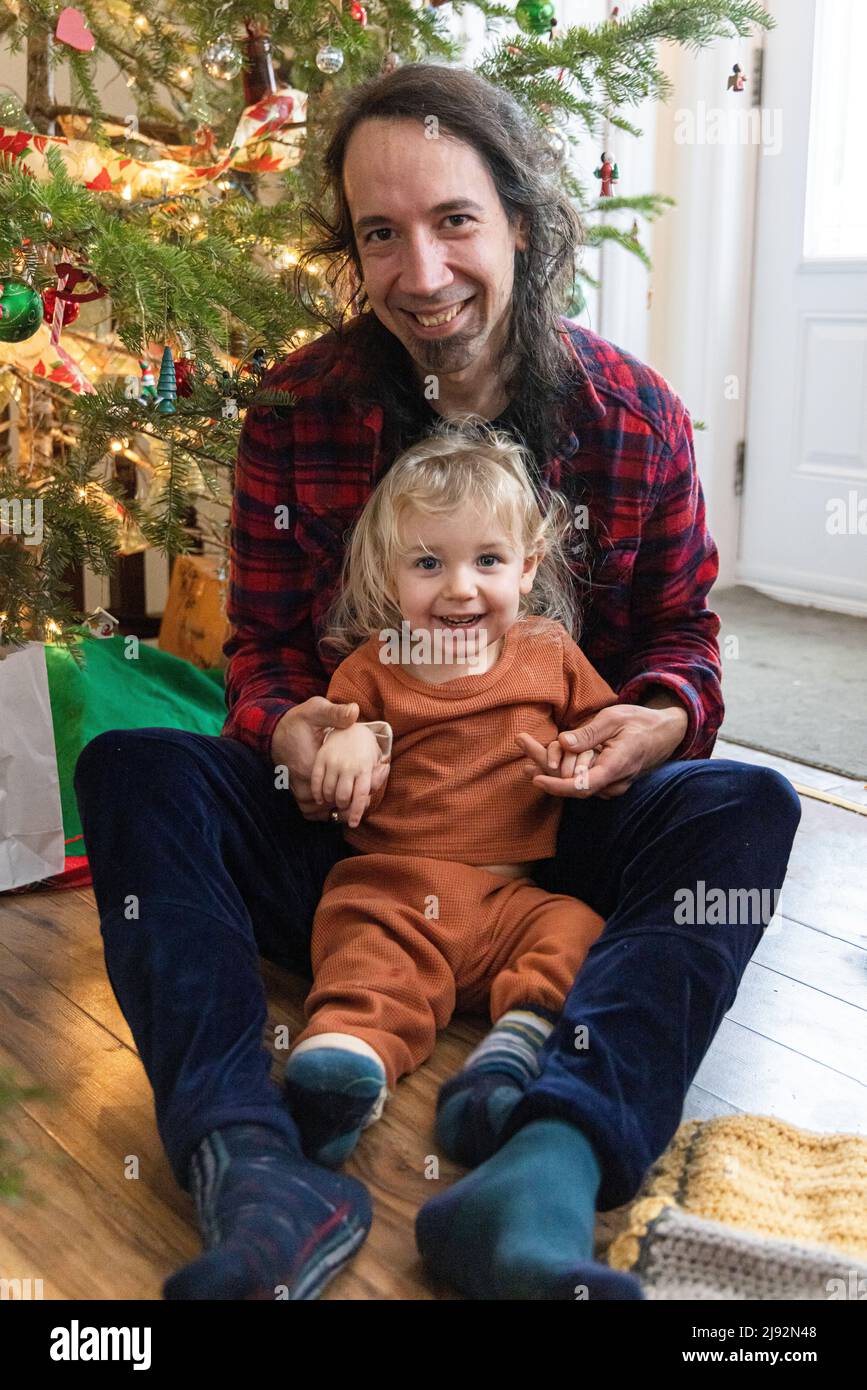 Dolce ritratto di famiglia di padre e figlio seduto da albero di natale al mattino di Natale. Con decorazioni sfocate sullo sfondo. Giovane famiglia caucasica. Foto Stock