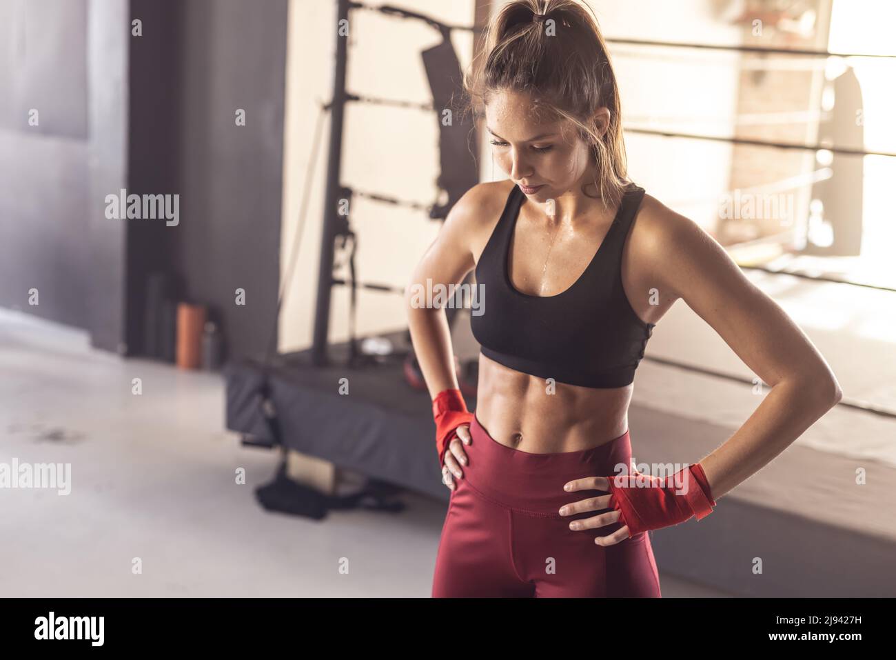 Boxer femminile giovane caucasico con involtini da boxe con le braccia akimbo in piedi nel centro benessere Foto Stock