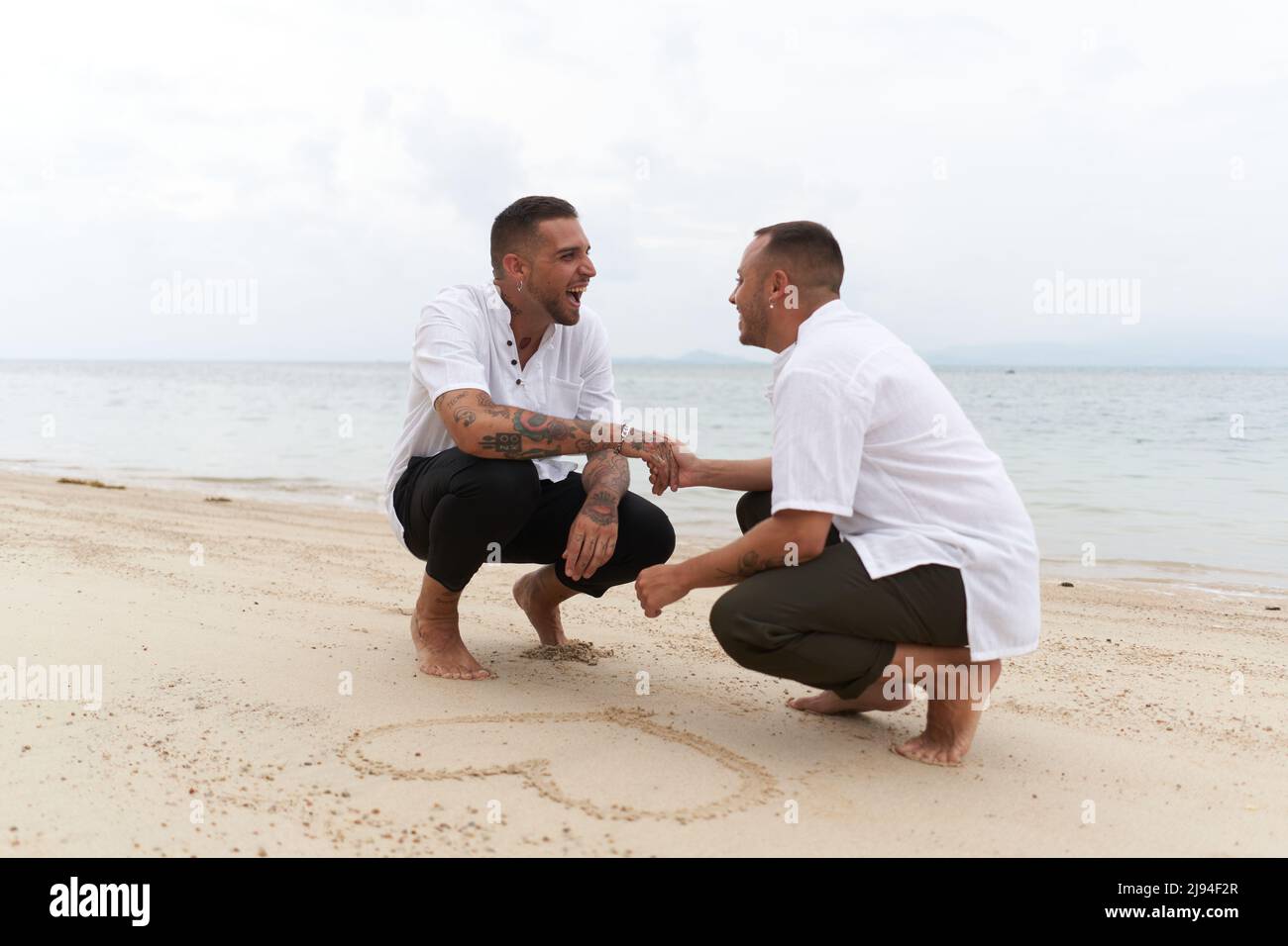Coppia gay che tiene le mani accanto ad un cuore disegnato nella sabbia su una spiaggia tropicale Foto Stock