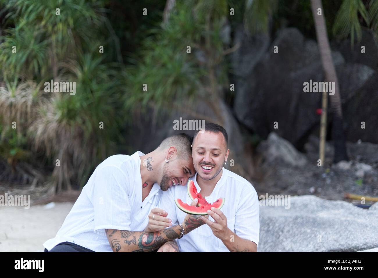 Uomo ridendo mentre si appoggia sul suo compagno mentre mangiava il cocomero su una spiaggia Foto Stock