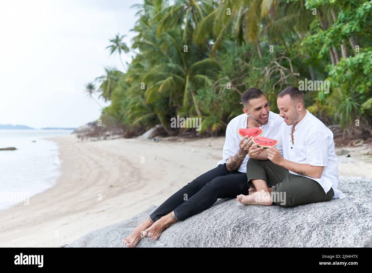 Coppia gay seduta su una spiaggia tropicale mentre mangiano il cocomero Foto Stock