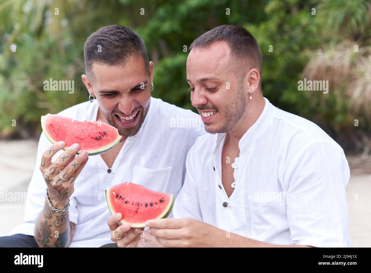 Due gay seduti su una spiaggia sorridendo e sorridendo un pezzo di cocomero Foto Stock