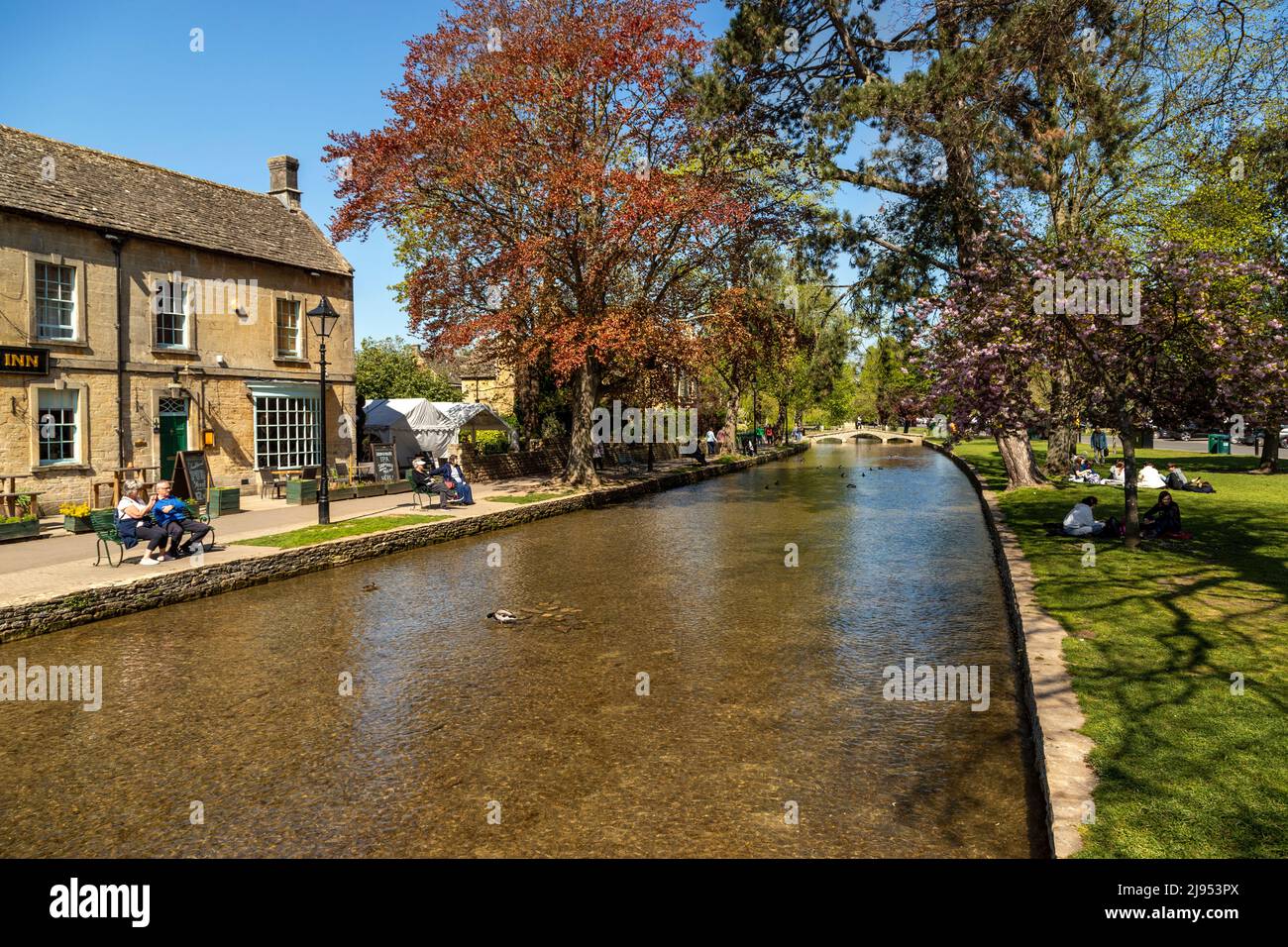 I visitatori possono godersi una giornata di sole in primavera a Bourton-on-the-Water lungo il fiume Windrush, una popolare destinazione del Cotswolds, Gloucestershire, Inghilterra, Regno Unito. Foto Stock