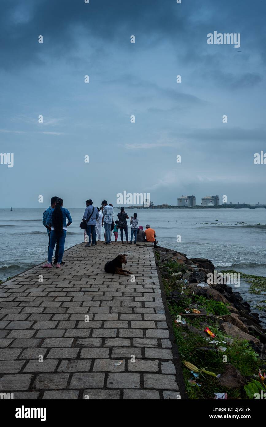 La gente si gode la brezza mattutina al passaggio pedonale di Fort Kochi con la foce del porto di Kochi e i carri armati di GNL sullo sfondo Foto Stock