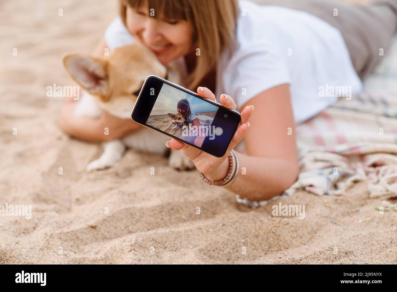 Donna anziana che scatta foto sul cellulare mentre riposa con il suo cane sulla spiaggia sabbiosa Foto Stock