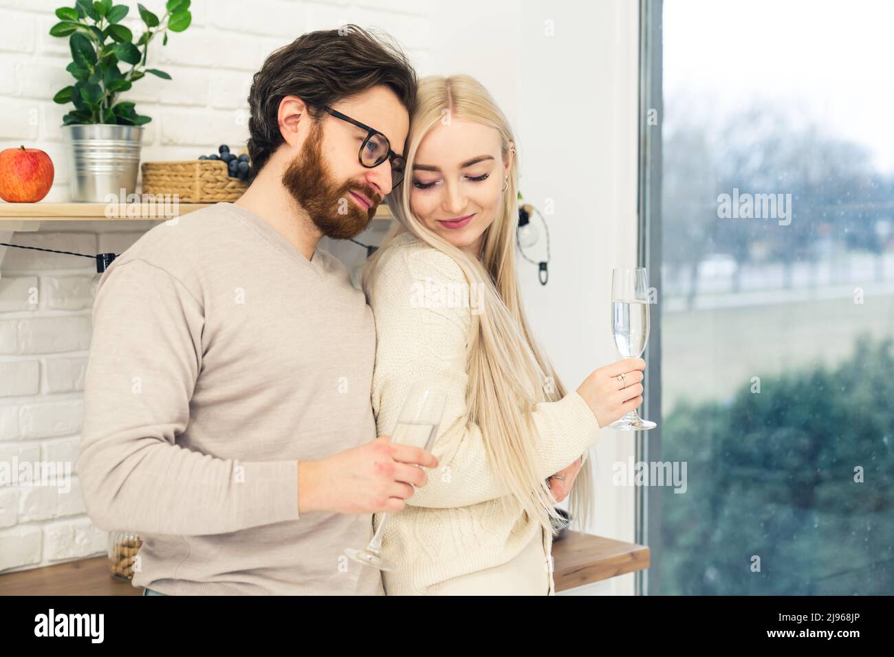 Bella sposata ragazzo e ragazza nel loro 20s in piedi vicino l'uno all'altro bere vino da bicchieri di vino in una cucina accanto a una grande finestra. Foto di alta qualità Foto Stock