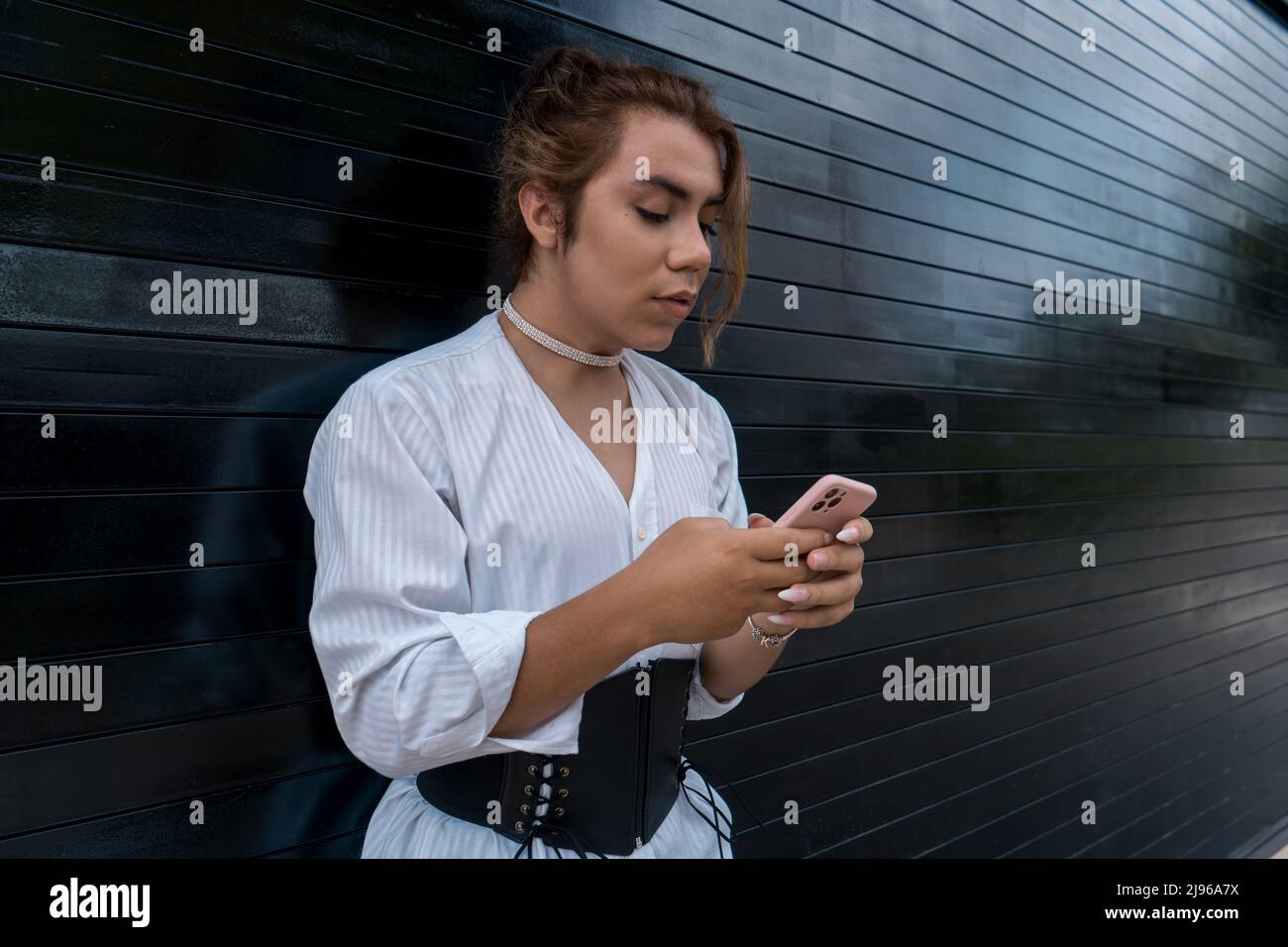 non binario utilizzando il telefono nelle sue mani chating appoggiandosi sul cancello nero in strada ricordando Foto Stock