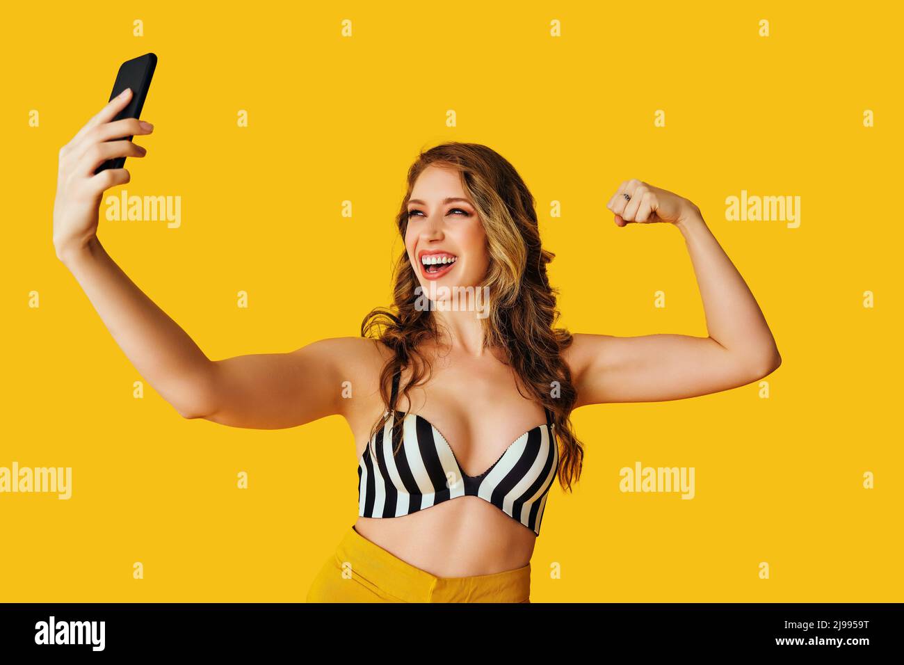 ritratto di bella giovane donna texting flirt bicep braccio muscolo selfie messaggio foto con smartphone su sfondo giallo studio Foto Stock
