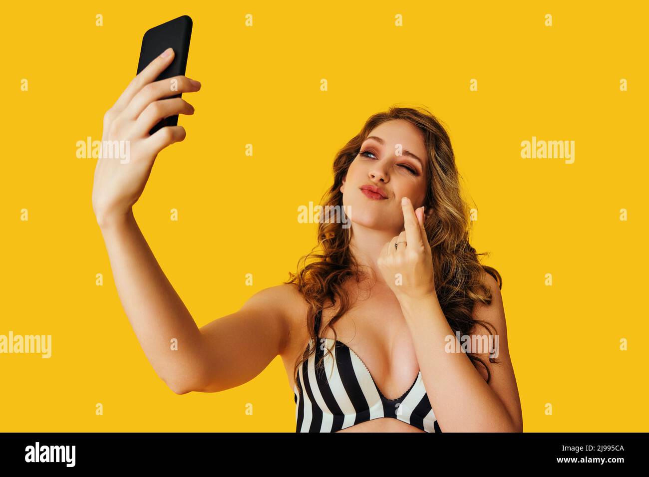 ritratto di bella giovane donna texting flirt selfie foto messaggio segreto denaro segno dita con smartphone su sfondo giallo studio Foto Stock