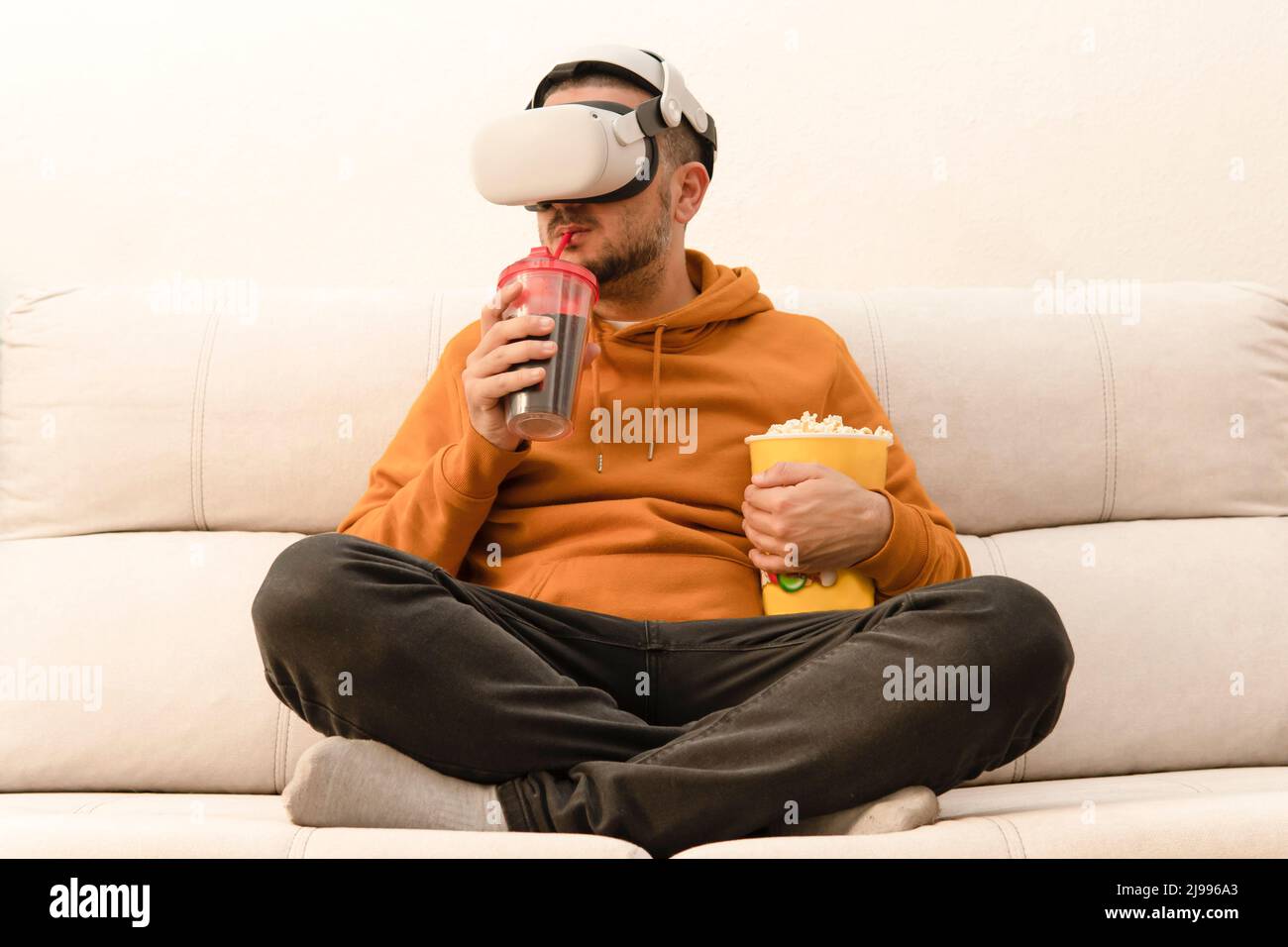 Uomo che guarda un film nei suoi occhiali di realtà virtuale bere soda e tenere popcorn mentre si siede sul divano Foto Stock