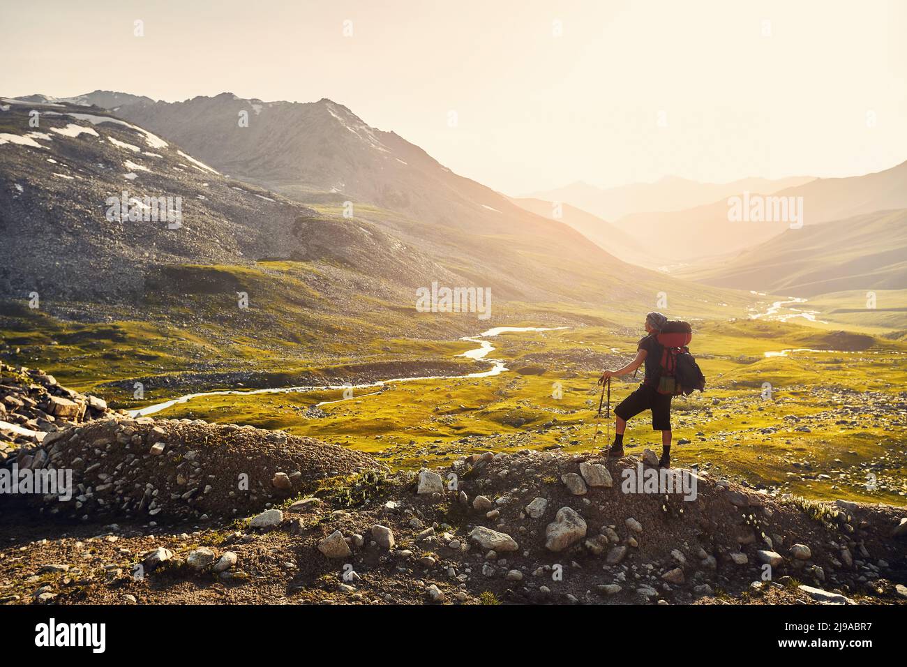 Silhouette di uomo Hiker uomo turistico con grande zaino in piedi sulla roccia contro il bellissimo tramonto e il fiume nella valle di montagna. All'aperto e trekking Foto Stock