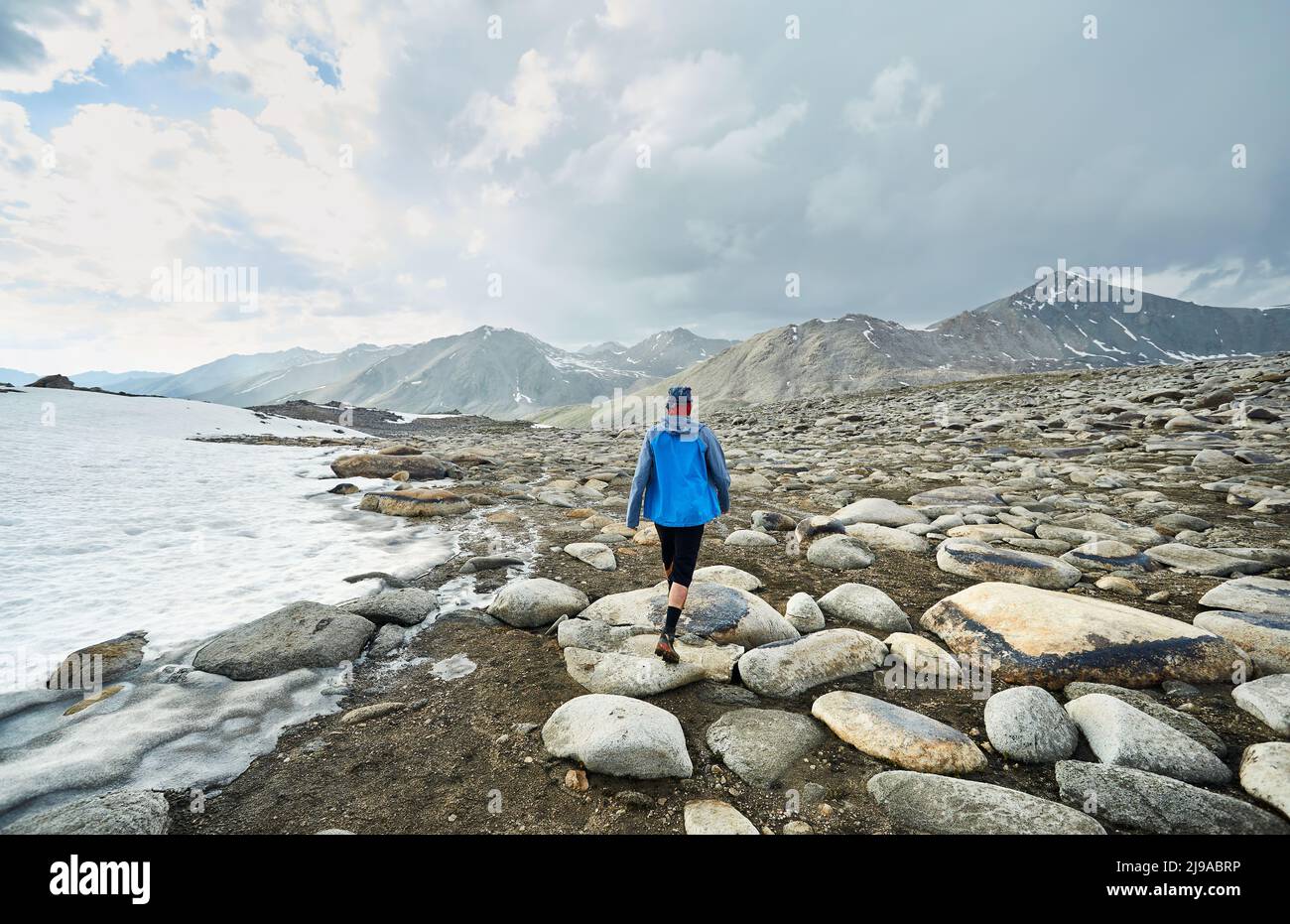 Uomo con giacca blu che cammina sulla valle rocciosa nelle belle montagne contro il cielo nuvoloso Foto Stock