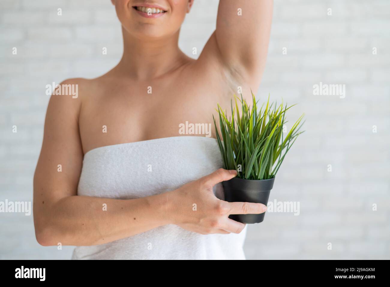 Donna sorridente in un asciugamano bianco tiene una pentola con una pianta. Ragazza irriconoscibile con l'aiuto di cespugli verdi imita la vegetazione dei capelli delle ascelle Foto Stock