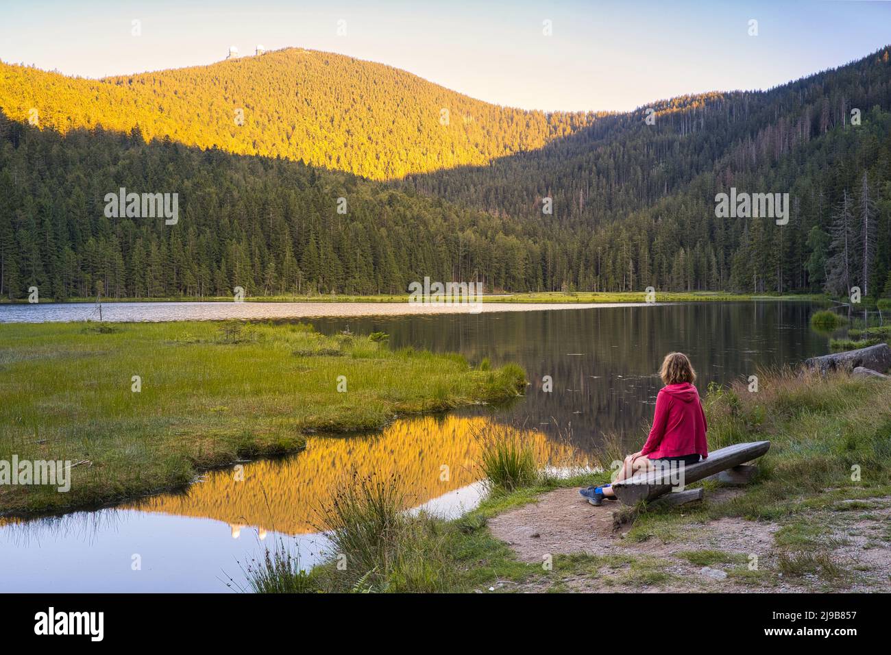 Una donna europea seduta su una panca di legno al bellissimo lago Kleiner Arbersee al tramonto. Vista sul monte Großer Arber con le sue due torri. Bavarese Fore Foto Stock