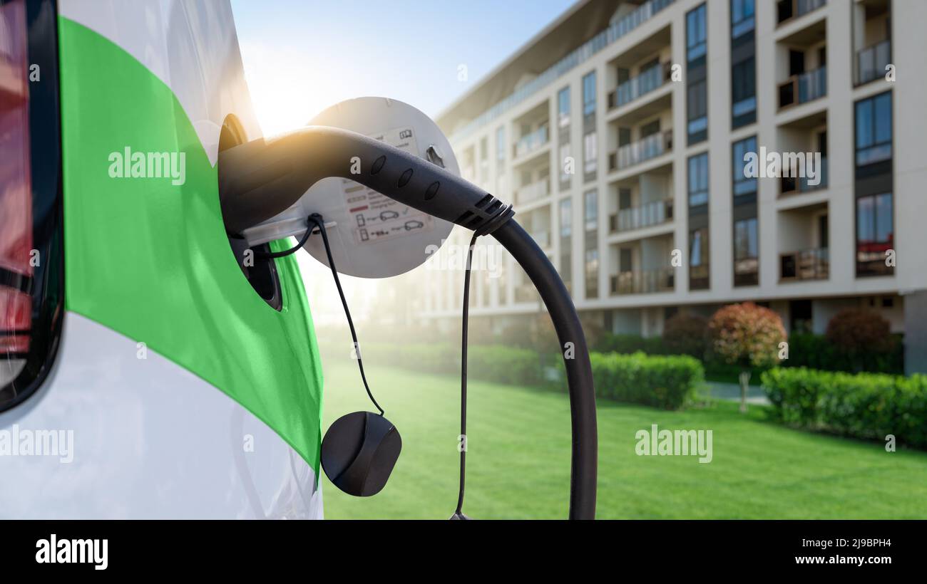 Primo piano dell'auto elettrica con un cavo di ricarica collegato sullo sfondo di una città ecologica verde Foto Stock