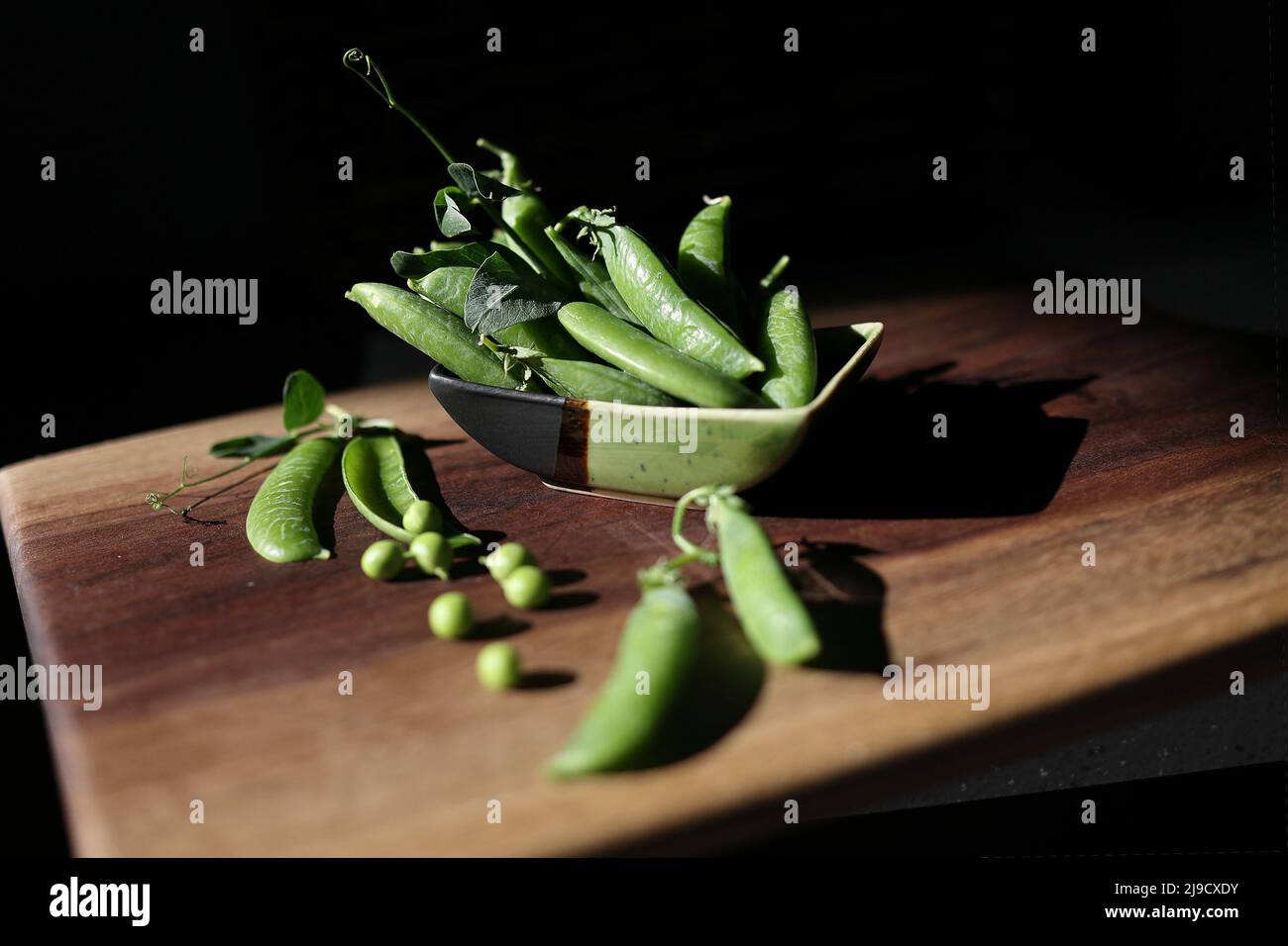 I deliziosi piselli verdi maturi si trovano in una piccola ciotola di ceramica e su un tavolo di legno. Sfondo scuro, stile rustico, luce solare intensa, messa a fuoco selettiva Foto Stock