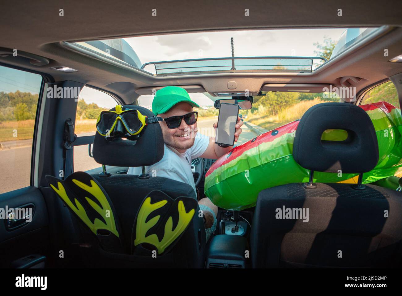 uomo guida felice auto pieno di roba di vacanza che tiene il telefono con schermo bianco Foto Stock