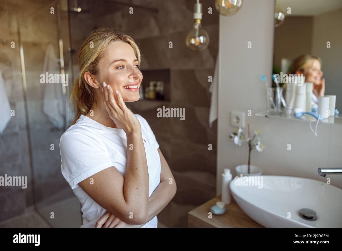 Sognante giovane bella donna bionda sorridente a specchio in bagno. Concetto di cura di sé. Foto Stock