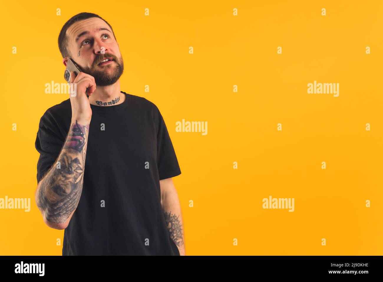 Tatuò l'uomo caucasico di mezza età che parla con il suo smartphone e guarda in alto. Concetto moderno di comunicazione. Studio medio girato su sfondo giallo. Foto di alta qualità Foto Stock