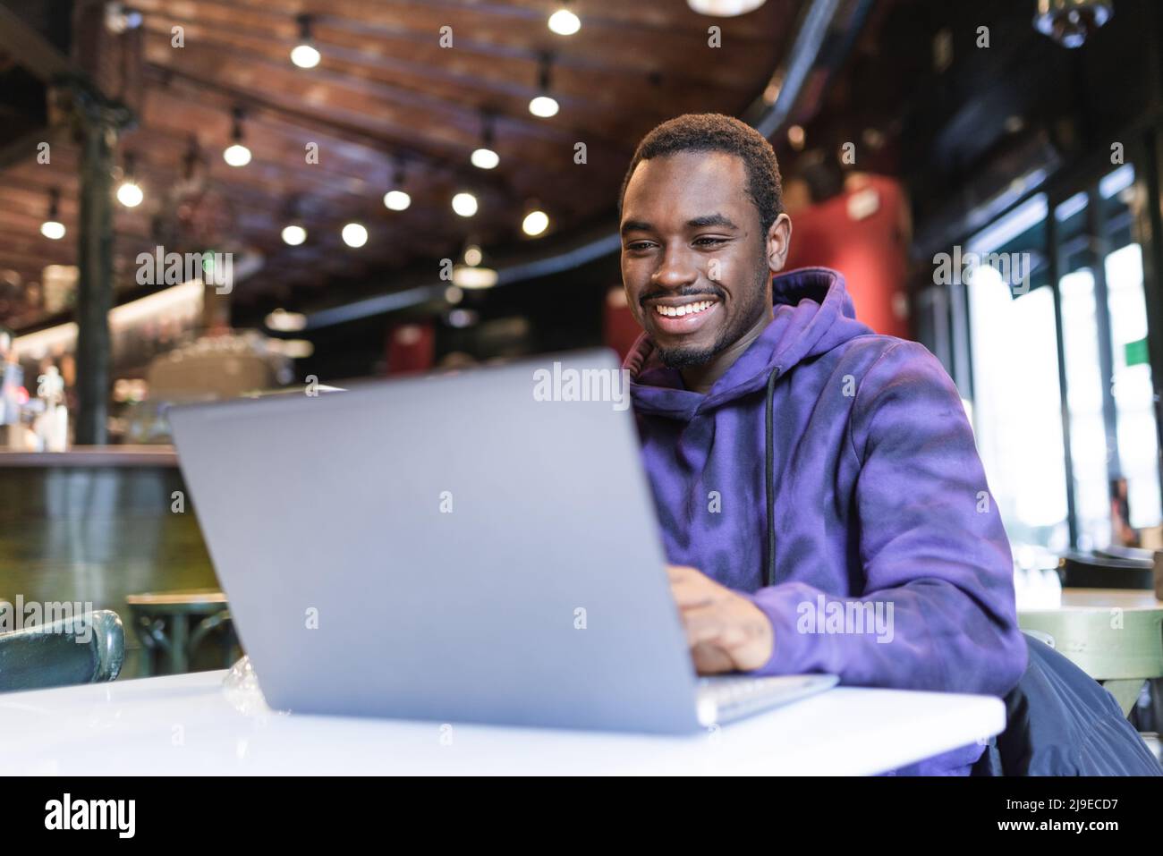 Positivo freelancer maschile afroamericano seduto al tavolo e digitando su netbook mentre si lavora a distanza in un moderno caffè Foto Stock