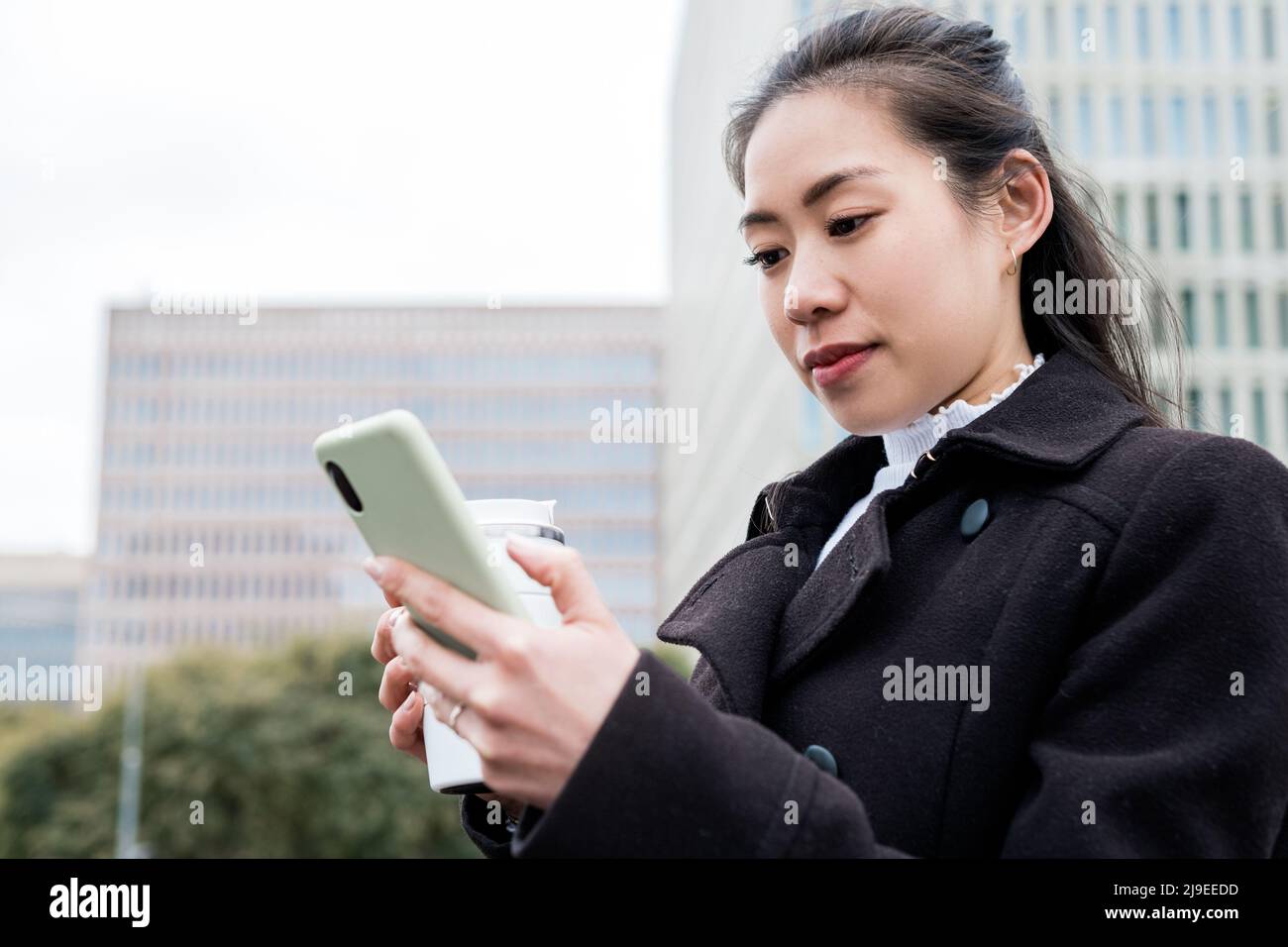 Basso angolo di donna d'affari cinese in cappotto nero con zero tazza di spreco di bevanda calda. Lei ir SMS sul cellulare durante la pausa caffè sulla strada della città Foto Stock