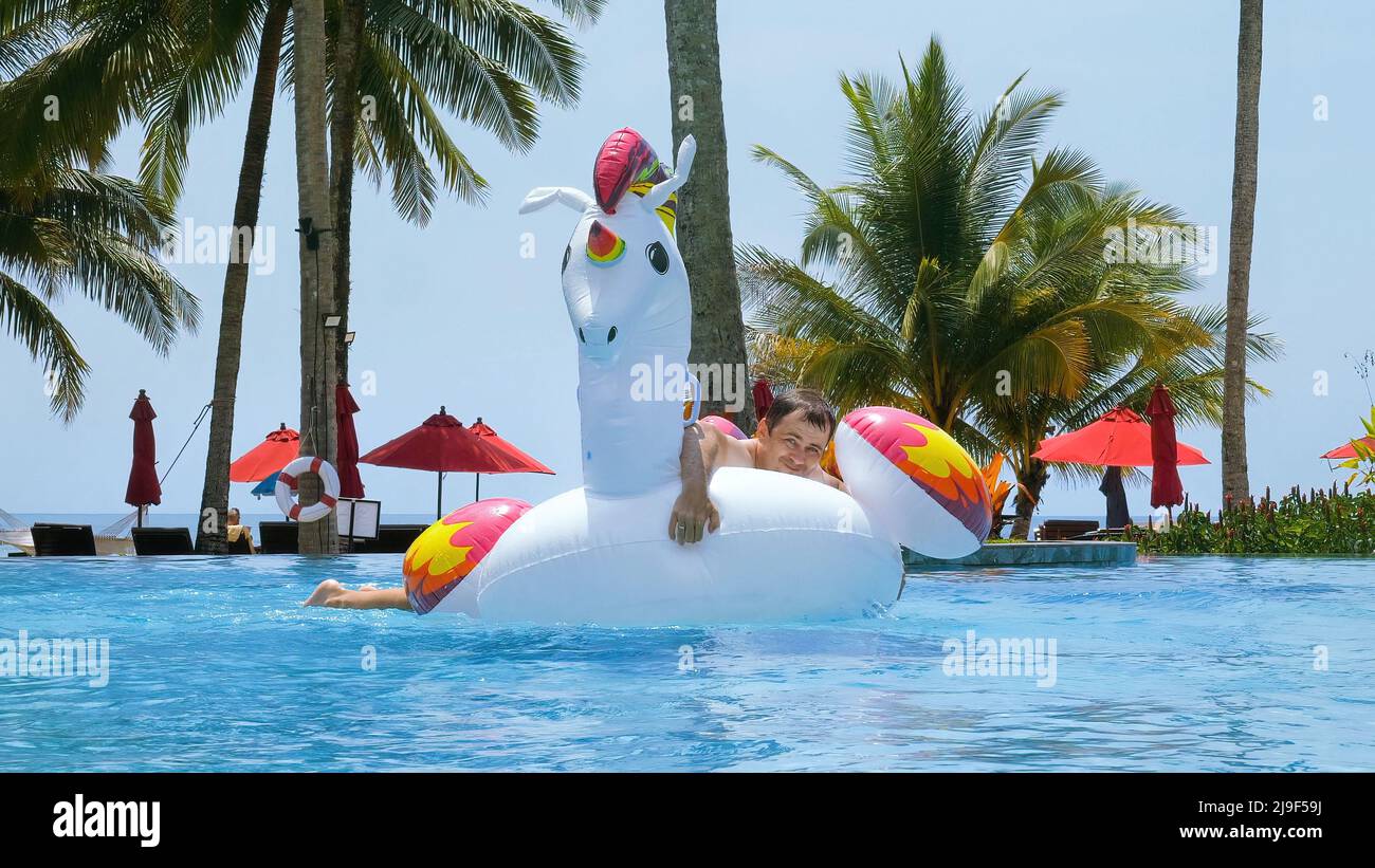 Felice uomo divertente con successo saltare su unicorno gonfiabile galleggiante in piscina in hotel tropicale per andare in giro. Pazzo turista maschile divertirsi su gigante Foto Stock