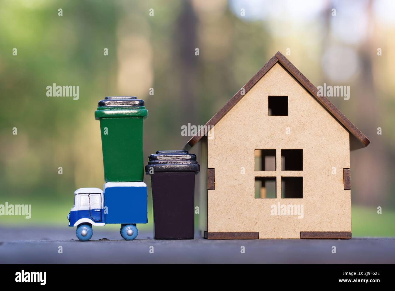 Carrello per rifiuti giocattolo con bidoni per rifiuti in piedi da un piccolo modello di casa in legno su uno sfondo di foresta. Raccolta di rifiuti in comunità rurali. Foto Stock