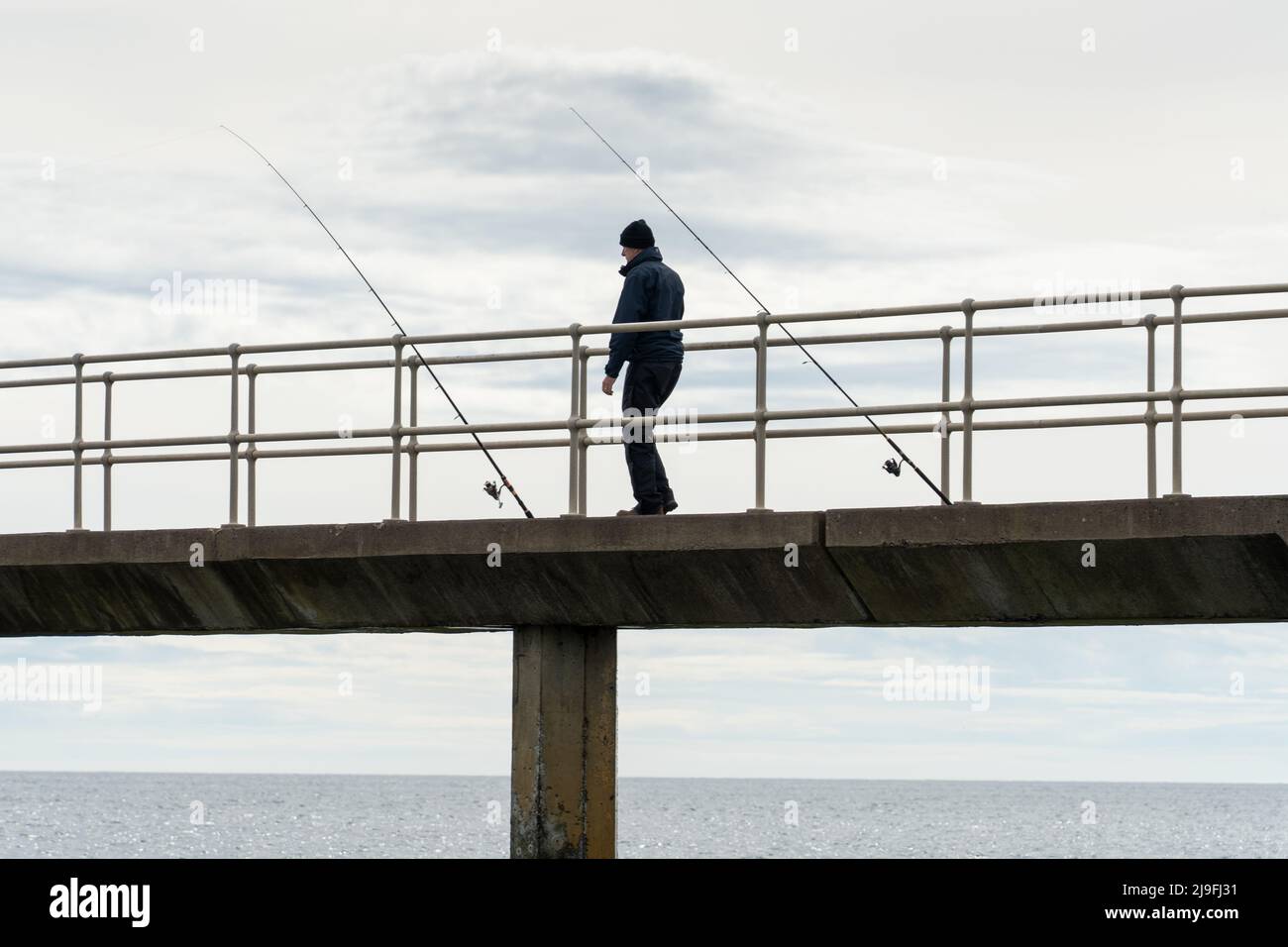 Un uomo si erge su un molo tra due canne da pesca in amble, Northumberland, Regno Unito. Foto Stock