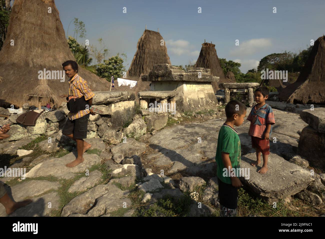 Un uomo che passa accanto ai bambini che giocano in uno spazio aperto dove si vedono tombe megalitiche nel villaggio tradizionale di Praijing a Tebara, Waikabubak, West Sumba, East Nusa Tenggara, Indonesia. Foto Stock