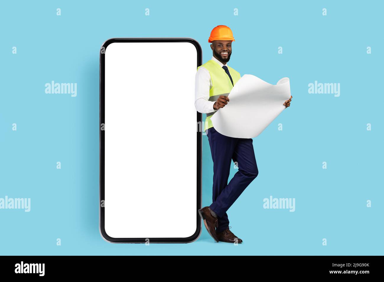 Black Smiling Construction Engineer con Blueprints Leaning su smartphone Big Blank Foto Stock