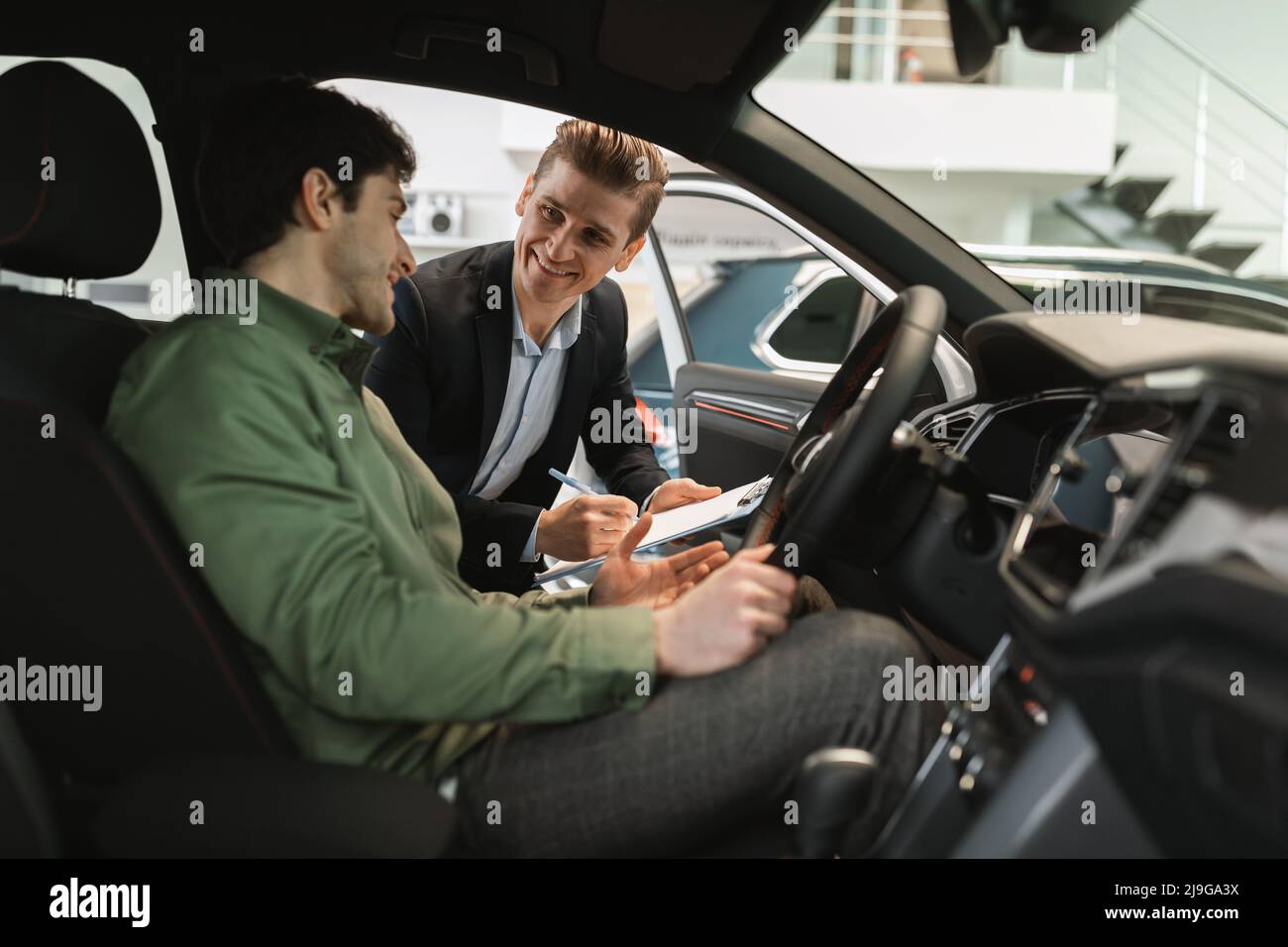 Giovane ragazzo caucasico che chiarisce i dettagli di acquisto dell'auto con il venditore, seduto all'interno del salone auto presso il centro concessionaria Foto Stock