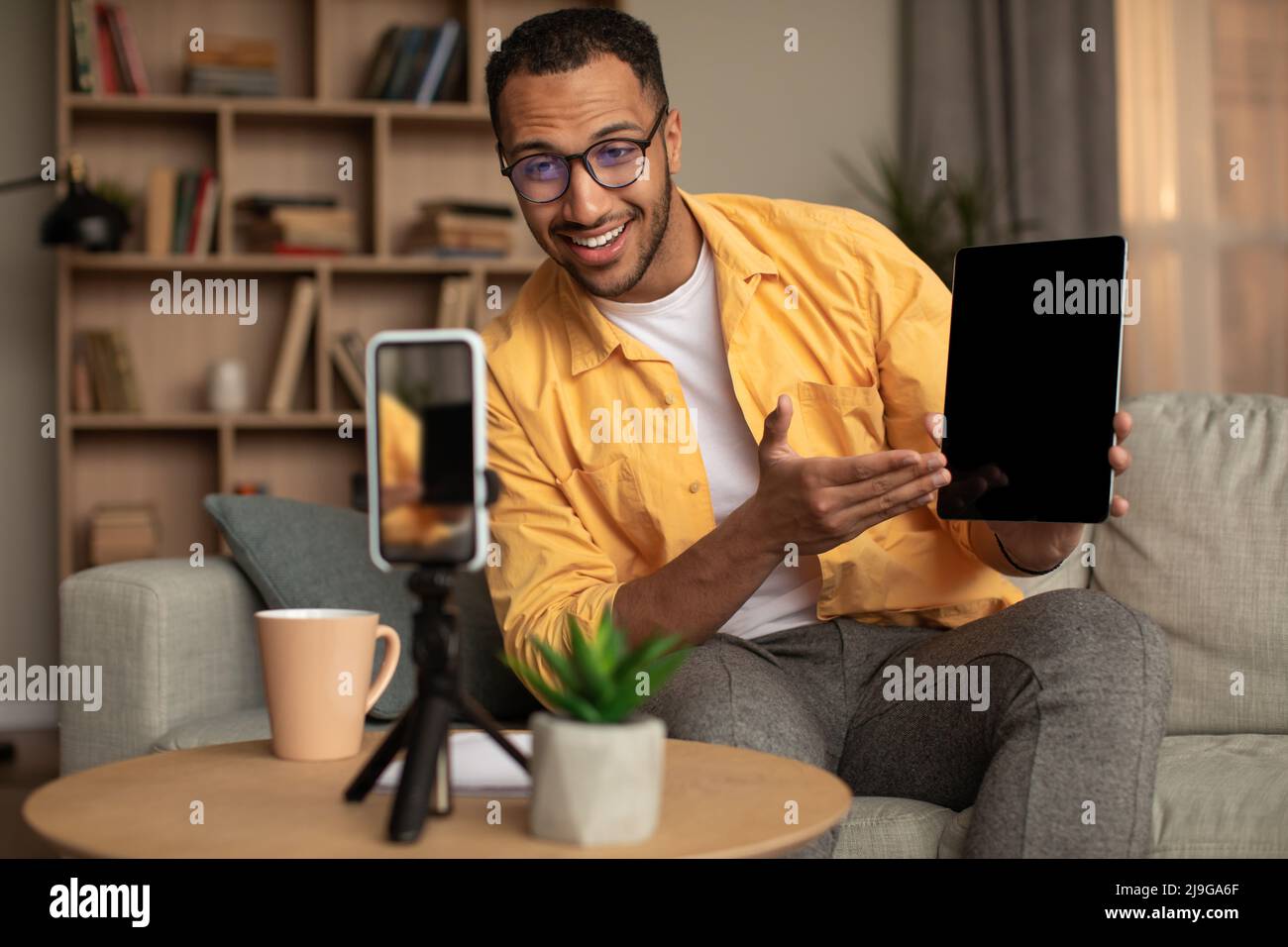 Allegro influencer nero maschio filma video recensione del nuovo tablet digitale su smartphone a casa, mockup Foto Stock