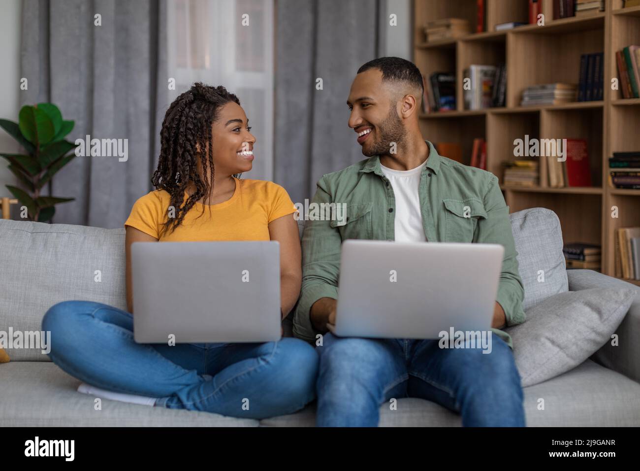 Giovane coppia nera che usa computer portatili, che guarda e sorride l'un l'altro, seduto sul divano e navigare in internet Foto Stock
