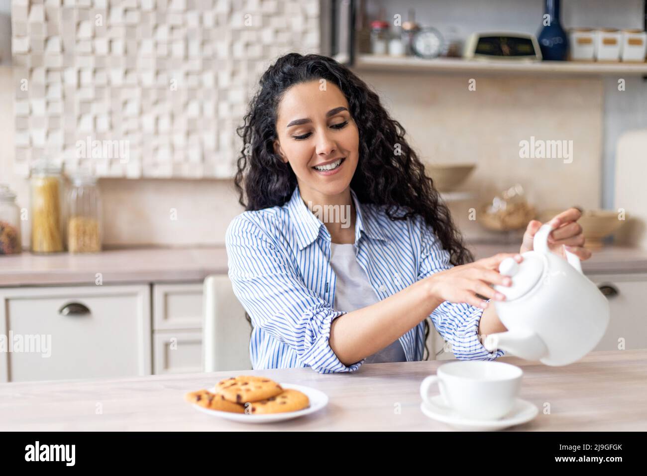 Positiva giovane donna latina che versa il tè in tazza, seduta al tavolo in cucina interna e godendo l'ora del mattino, spazio libero Foto Stock