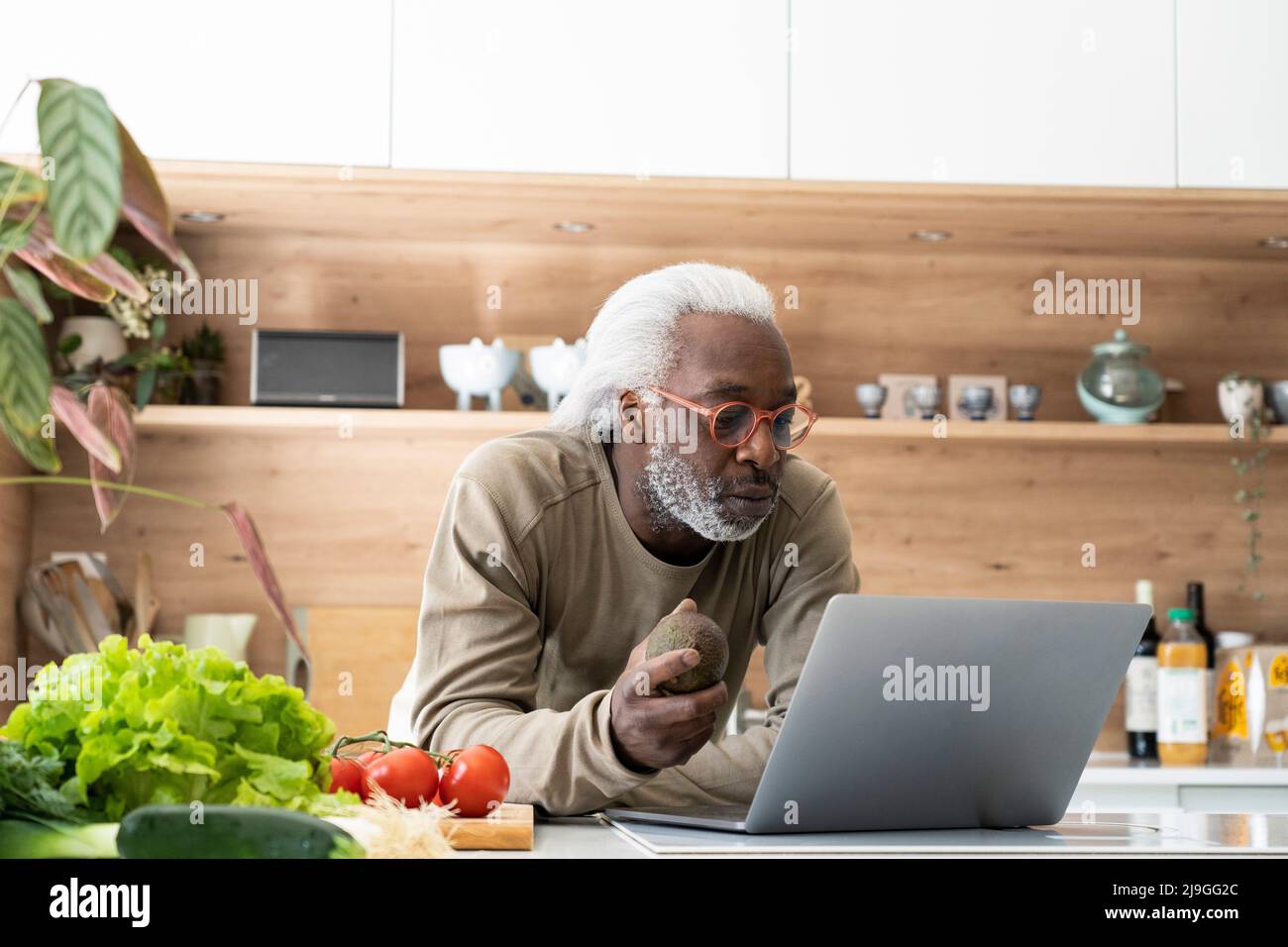 Uomo anziano che guarda la ricetta sul laptop mentre si appoggia sul piano di lavoro della cucina Foto Stock