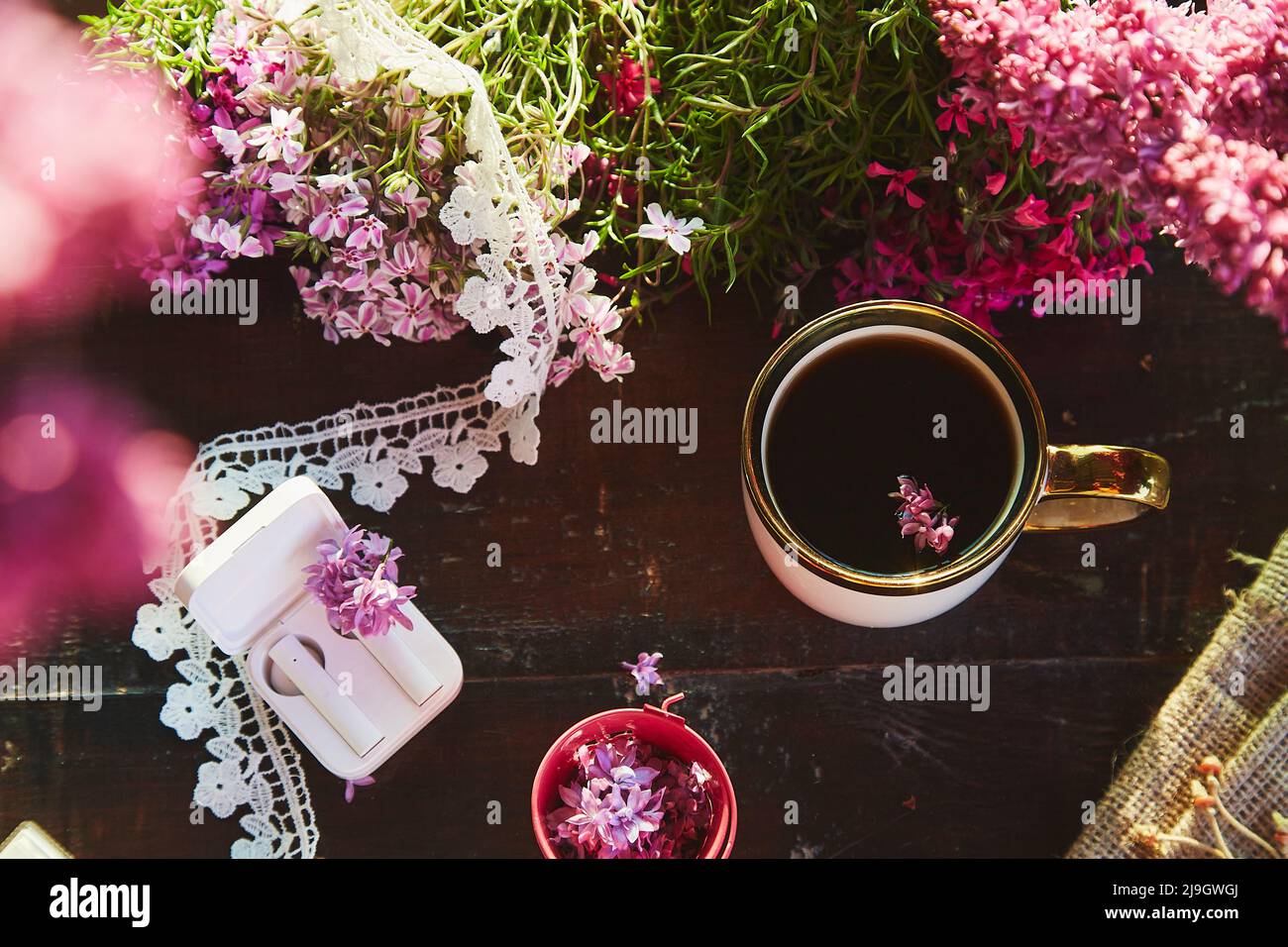 Stile di vita estetico piatto, cuffie senza fili, fiori lilla decorazioni e tazza di caffè. Ascolto di musica o audiolibro, webinar o chat online, conversazione telefonica in giardino Foto Stock