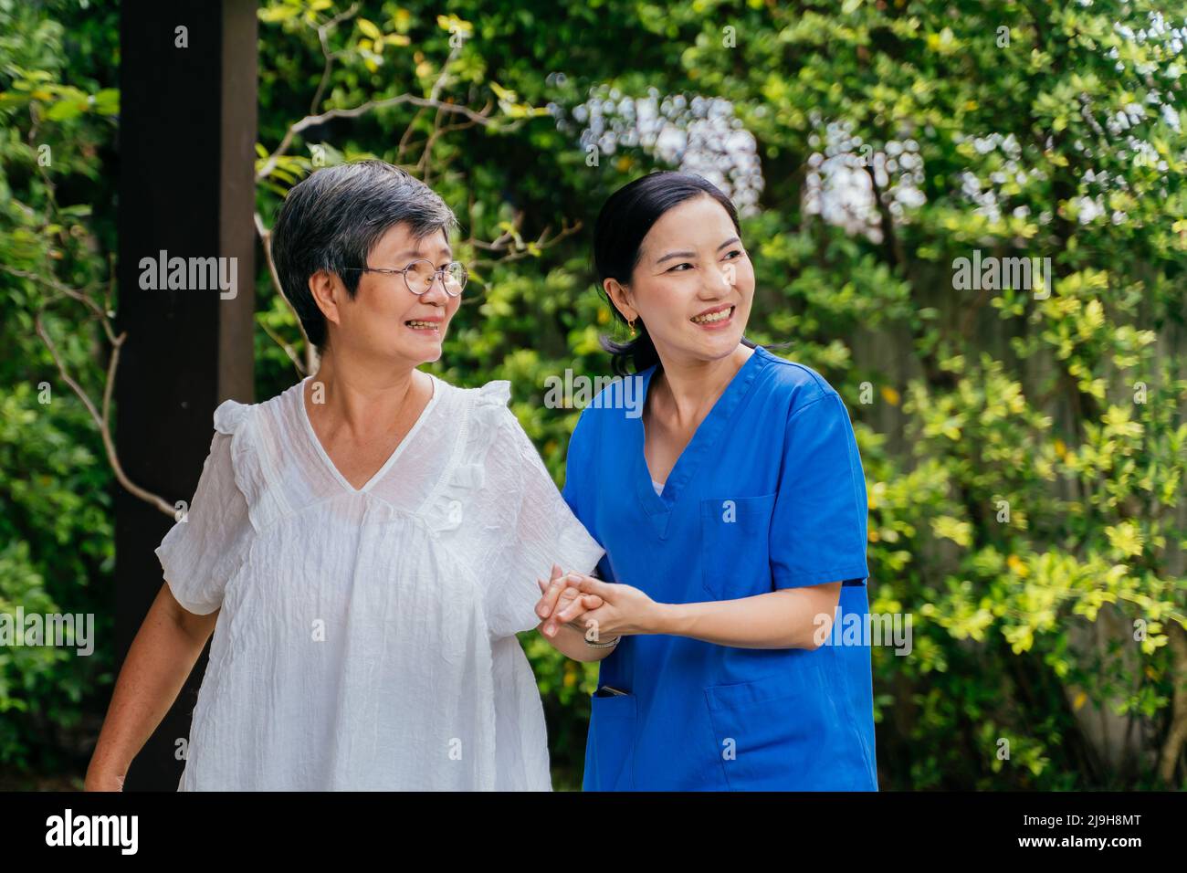 Donna felice caregiver in scrub blu che tiene la mano della donna anziana e guardando via con il sorriso mentre cammina vicino alberi lussureggianti in giardino insieme Foto Stock