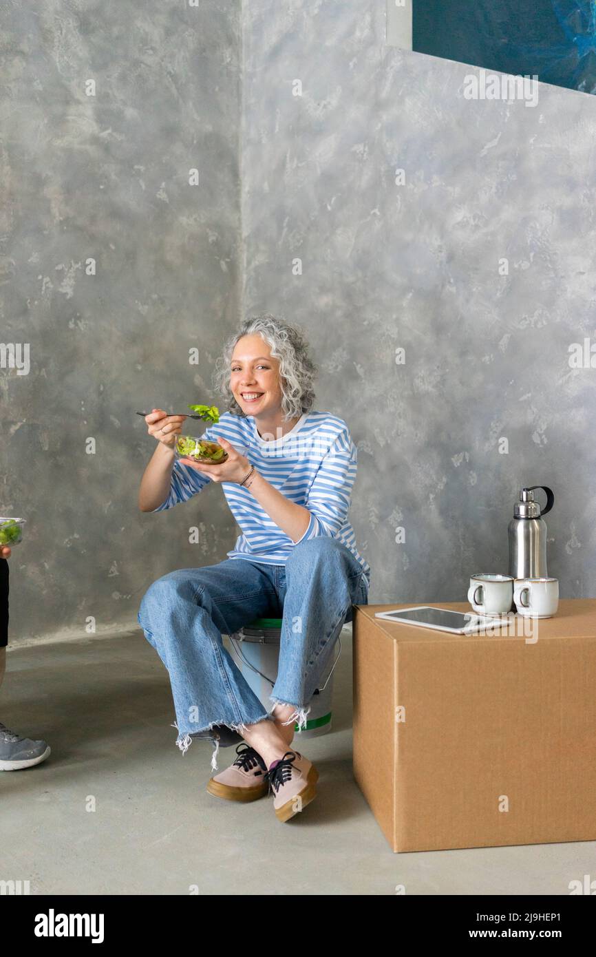 Donna sorridente che mangia il pranzo di fronte al muro grigio Foto Stock