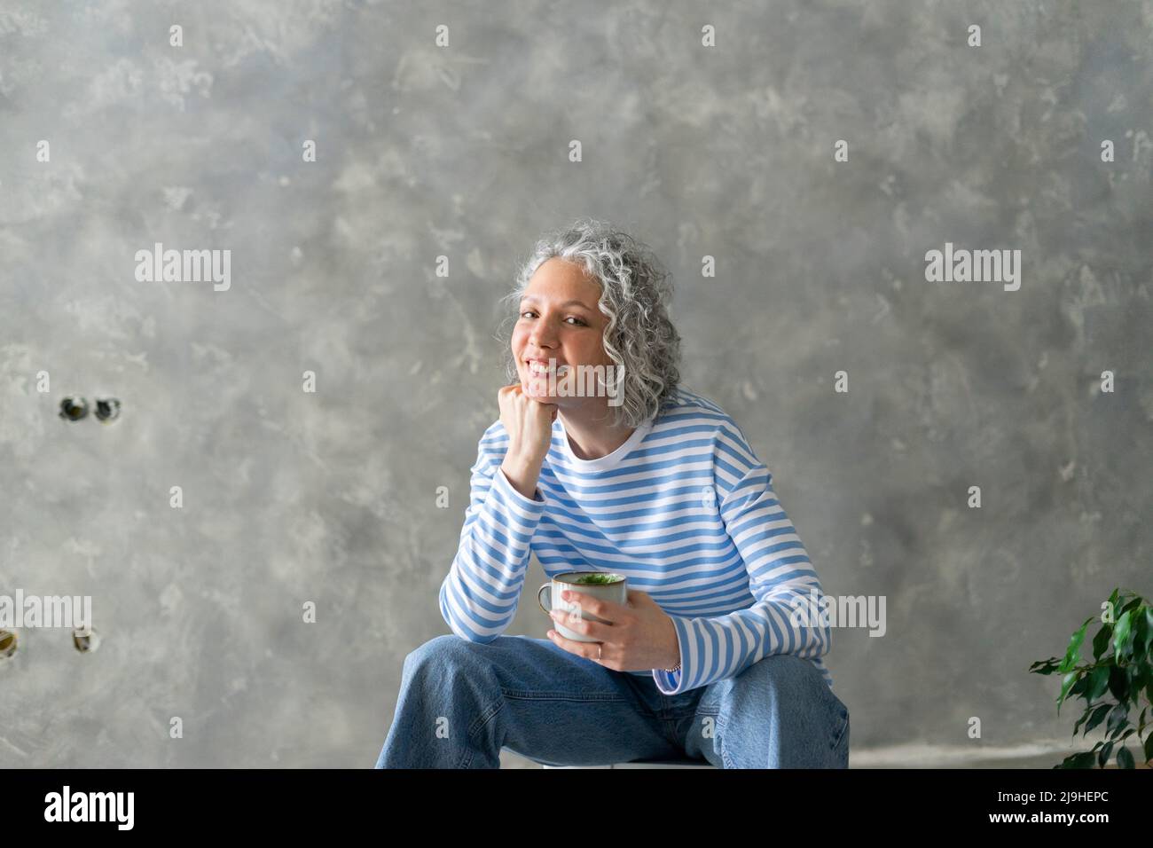 Donna felice con mano sul mento che tiene la tazza seduta davanti al muro Foto Stock