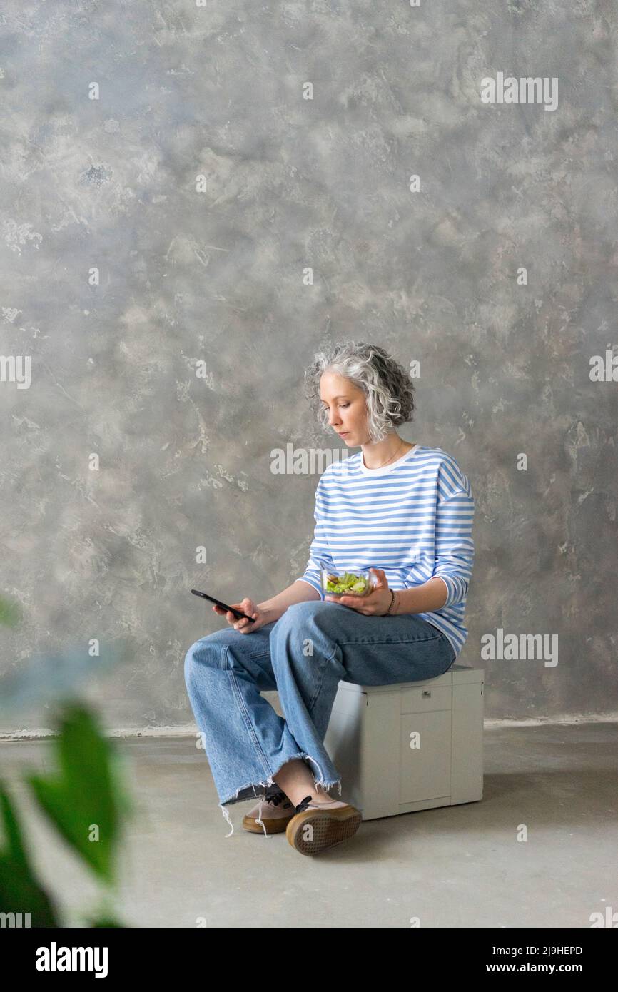 Donna che usa lo smartphone seduto sul box con pranzo davanti alla parete grigia Foto Stock