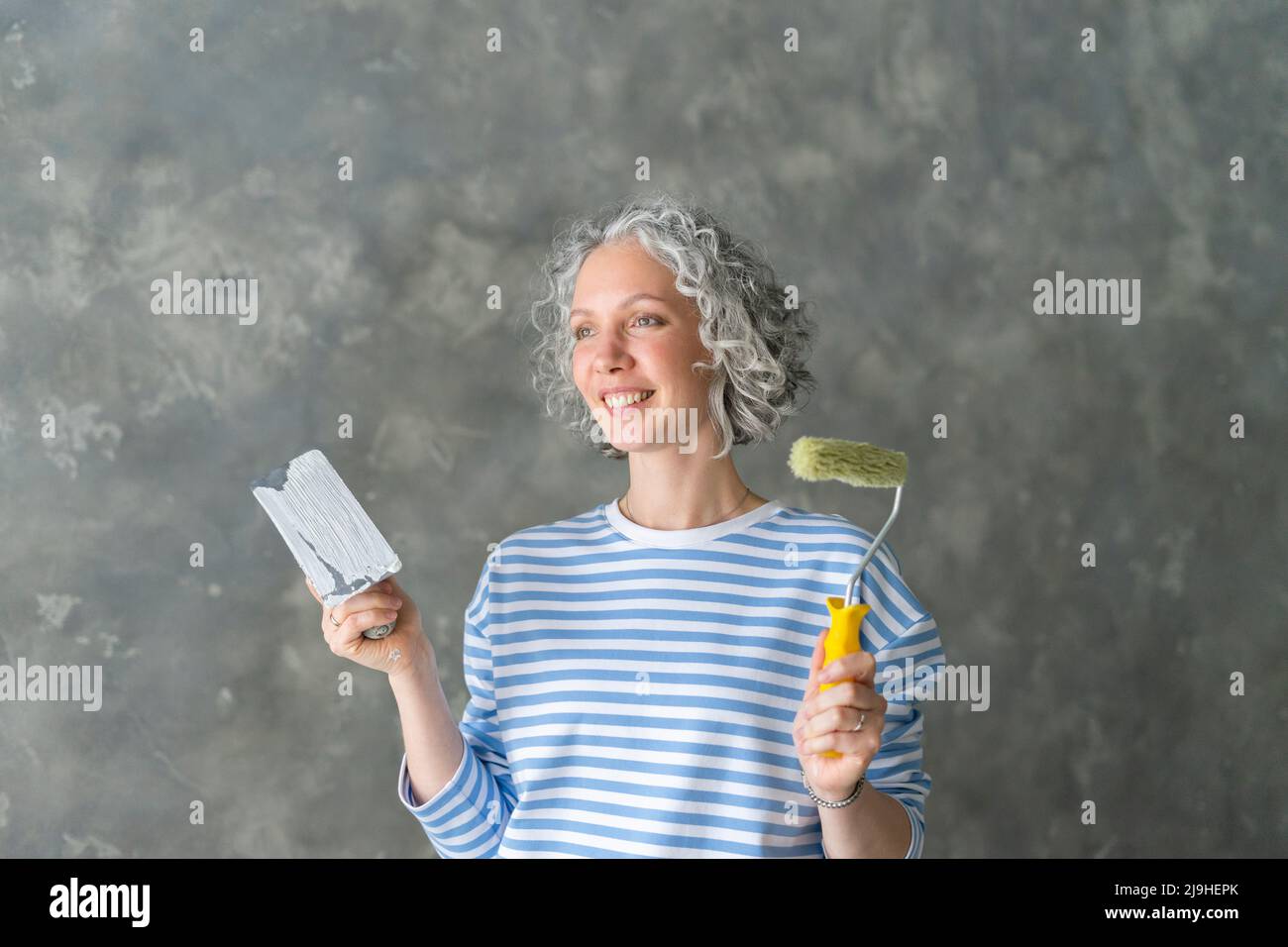 Donna sorridente con rullo in vernice e attrezzatura da lavoro in piedi davanti al muro Foto Stock