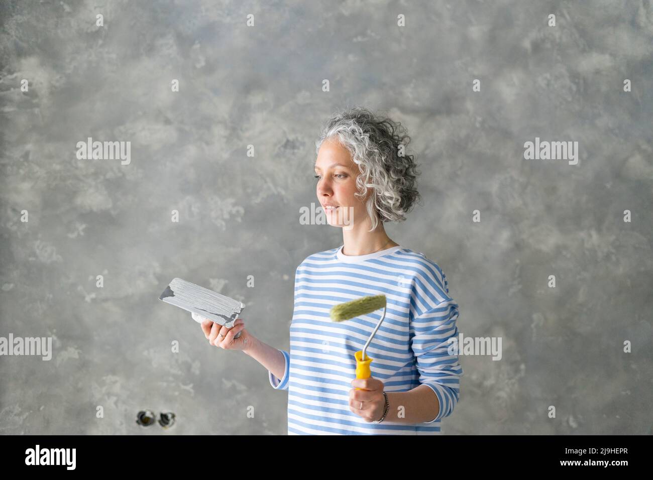Donna con rullo per pittura e attrezzatura in piedi davanti alla parete grigia Foto Stock