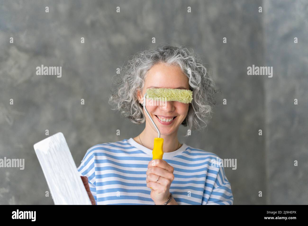 Donna sorridente che copre gli occhi con rullo di vernice davanti alla parete Foto Stock